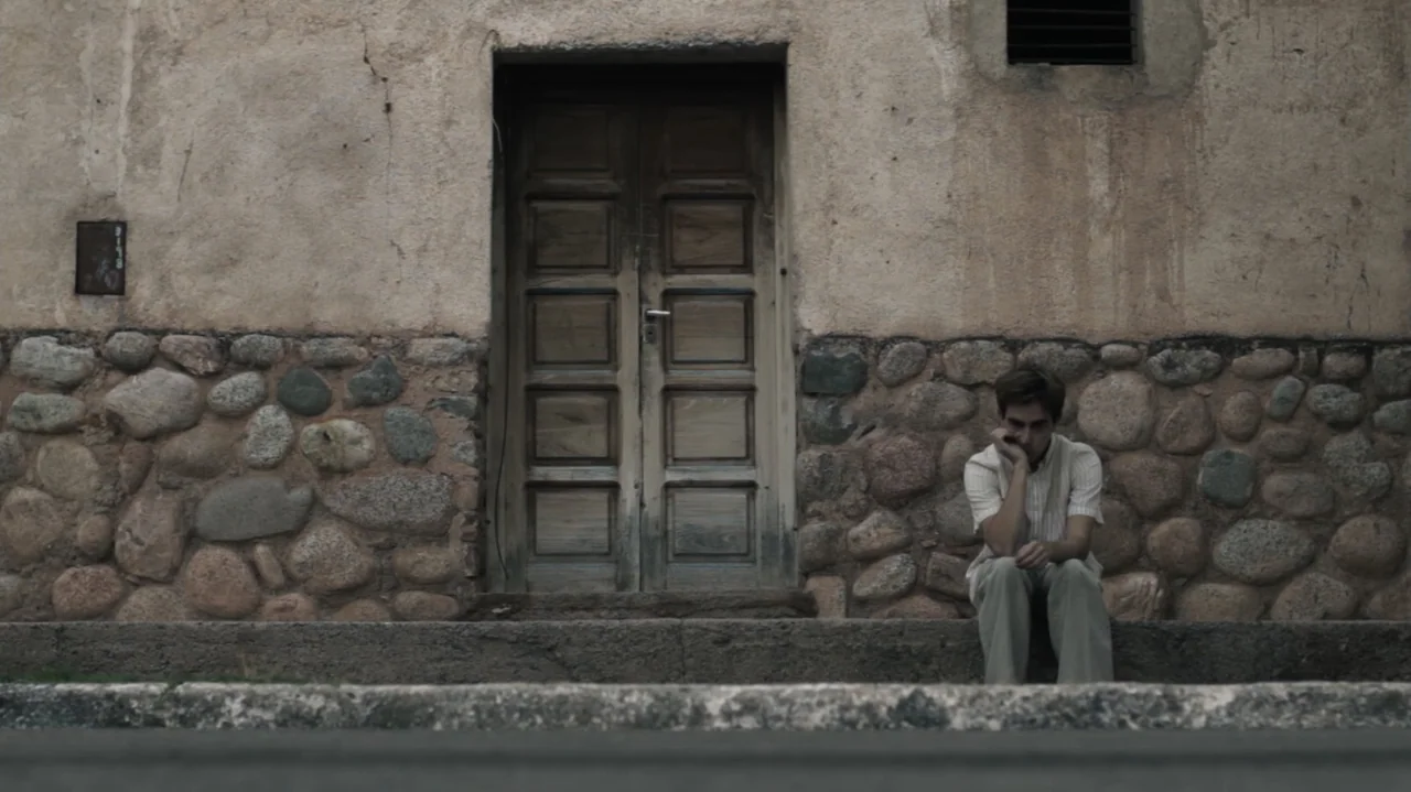 Still Life (2013) movie still: medium — A man sits on a low stone ledge in front of an old wall with a wooden door. He is positioned to the right,…