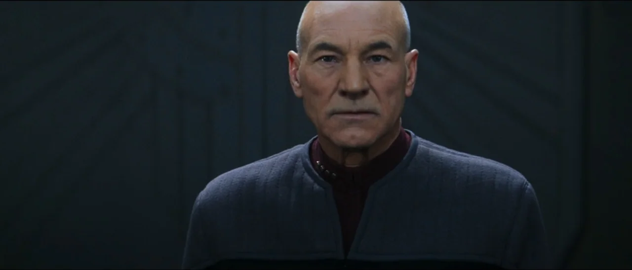 Star Trek: Nemesis (2002) movie still: medium — A dark, minimalist interior with subtle geometric paneling on the walls. A bald man faces the camera…