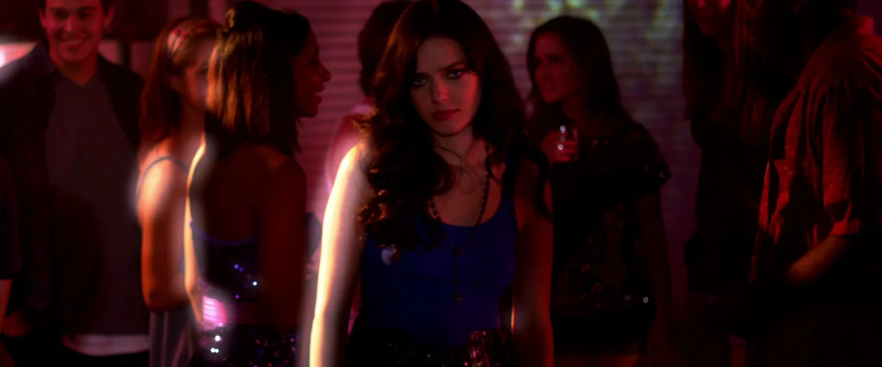 Kaboom (2010) movie still: medium — A dimly lit room filled with partygoers standing close together. A woman in a blue top stands in the center…