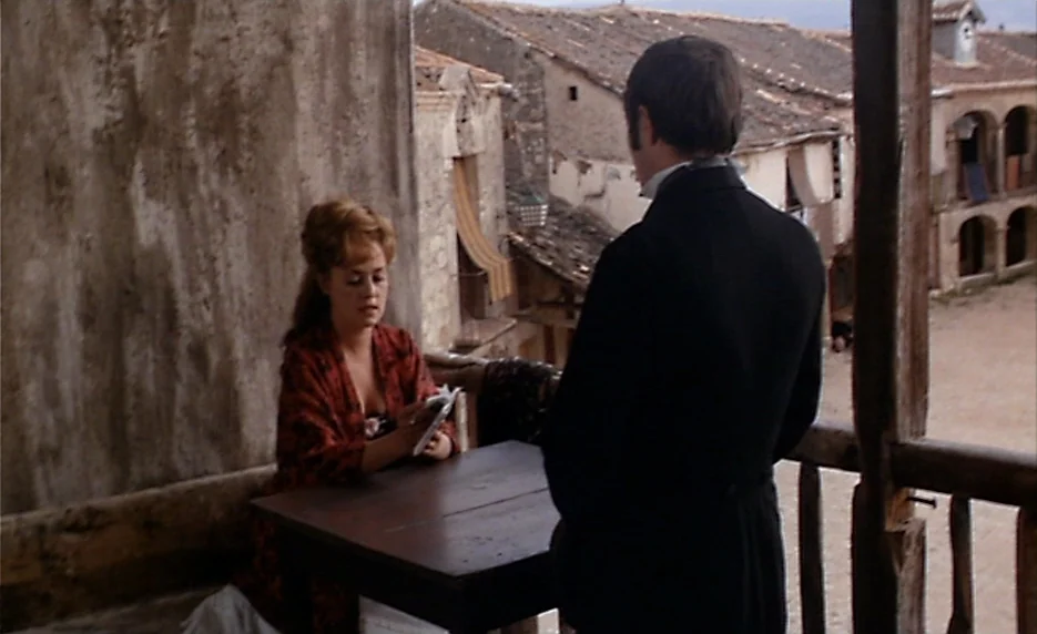The Immortal Story (1968) movie still: medium — A rustic balcony overlooks a dusty town square with traditional stone buildings. A man in a formal…