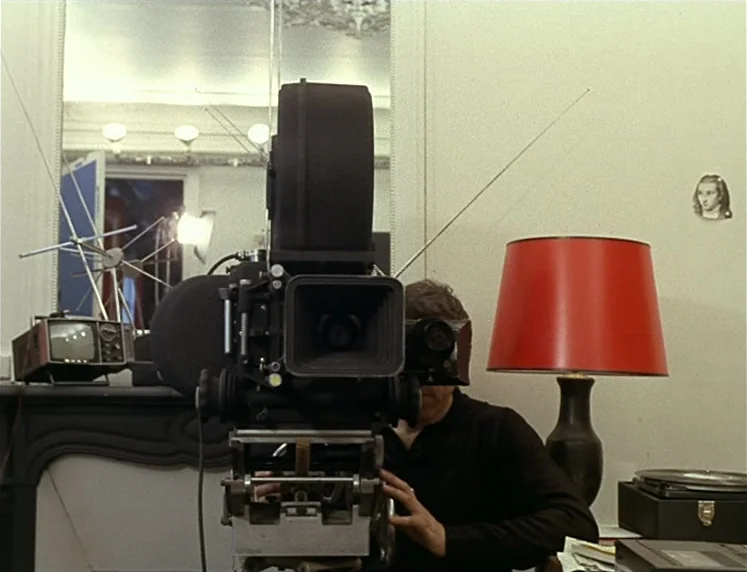 La Chinoise (1967) movie still: medium — A man sits behind a large professional movie camera in a sparsely furnished room. A bright red table lamp…