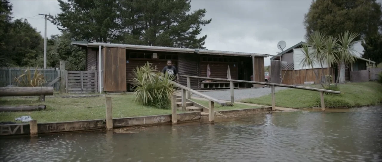 Bludgeon (2018) movie still: wide — A wooden building sits on the edge of a calm, murky lake with grass. A man walks down a wooden ramp toward the…