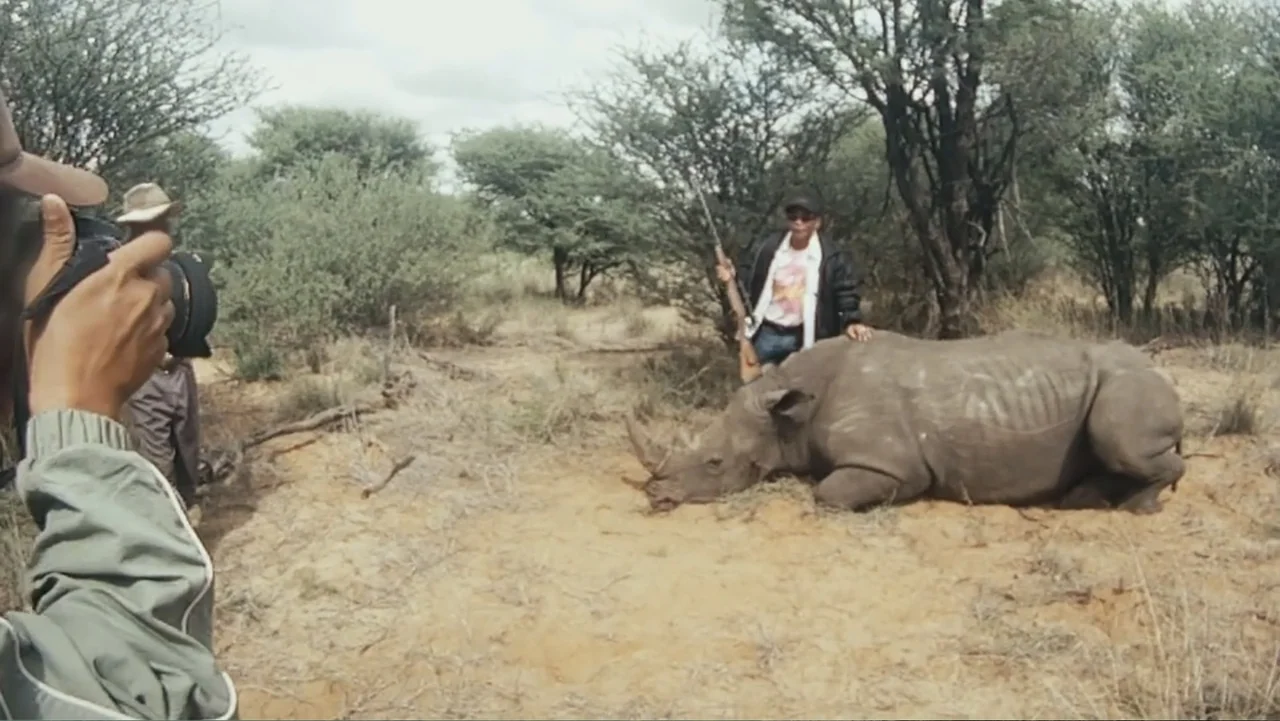 Bloody Trophy (2025) movie still: medium — A dry savanna landscape features scattered trees and sandy ground under an overcast sky. A man in a dark…