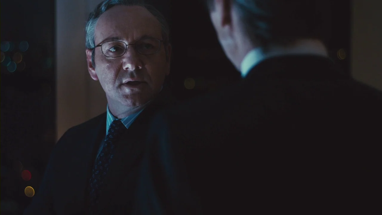 Margin Call (2011) movie still: medium — A dark office at night with city lights visible as bokeh in the distance. A man in a suit looks towards an…