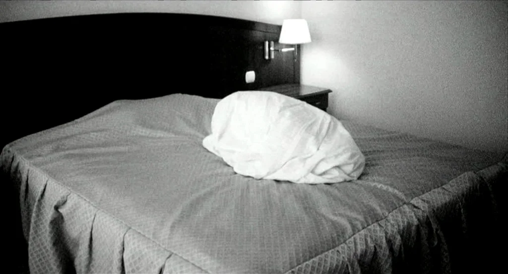 Container (2006) movie still: medium — A dimly lit hotel room features a large bed with a dark headboard. A single white blanket covers an…