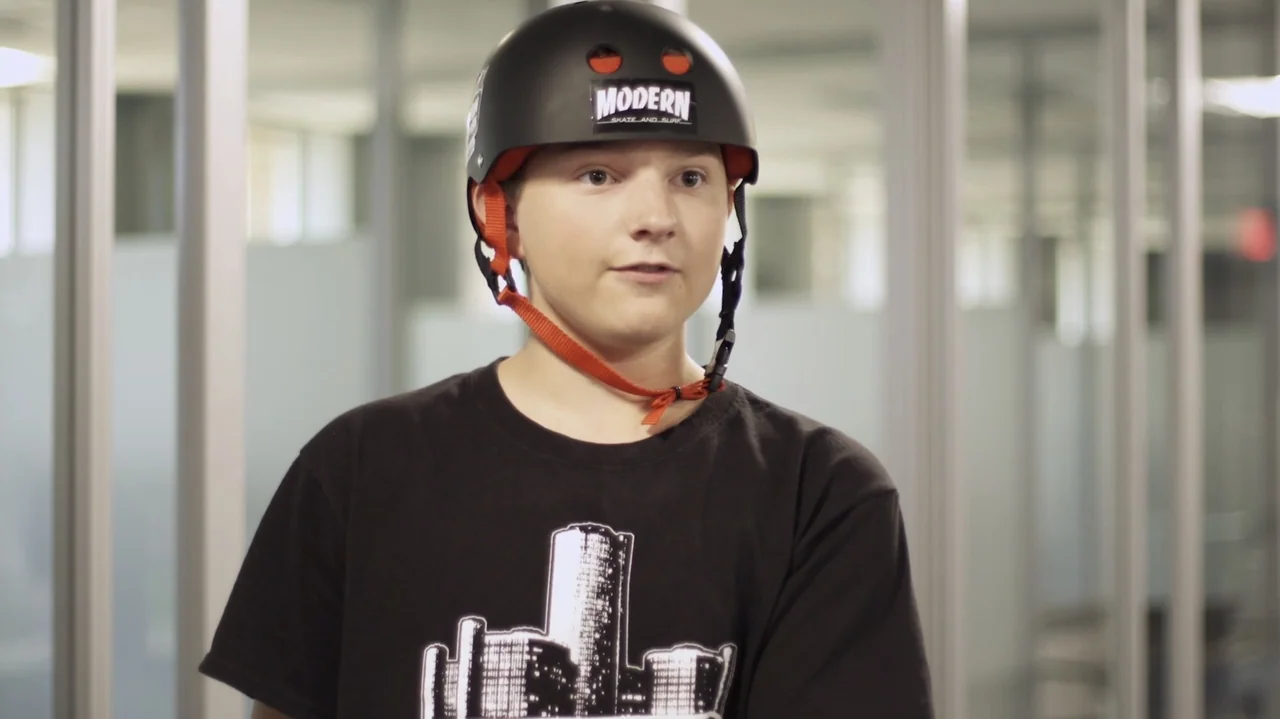 Sk8 Dawg (2018) movie still: medium — A sterile office interior features glass partition walls and a blurred white background. A teenager wearing a…