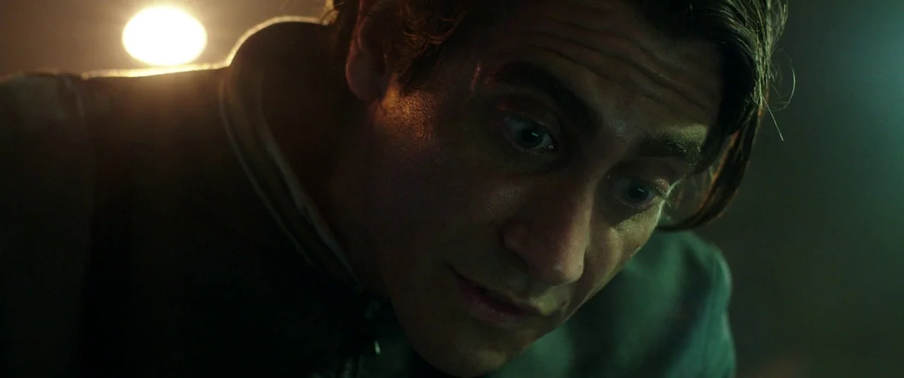 Nightcrawler (2014) movie still: closeup — A dimly lit room provides no background detail due to deep shadows. A man leans forward while looking…