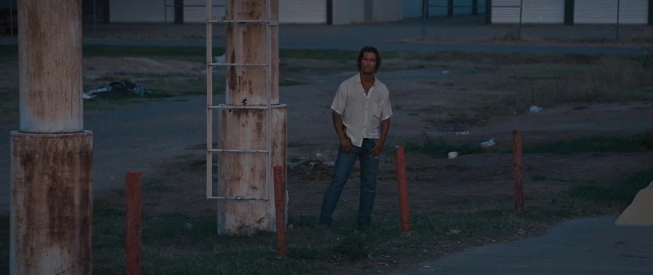 Mud (2012) movie still: wide — A gravel lot at dusk sits before a row of corrugated metal storage units. A man wearing a cream shirt and jeans…