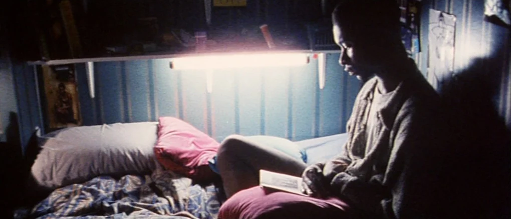 Wonderland (1999) movie still: medium — A small room features blue vertical-striped walls and a bed with rumpled blankets. A young man sits on the…