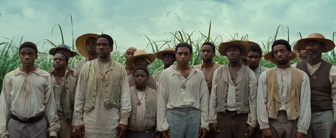 12 Years a Slave (2013) movie still: medium — A group of laborers stand in a dense field of tall green sugarcane. The sky is overcast and the men…