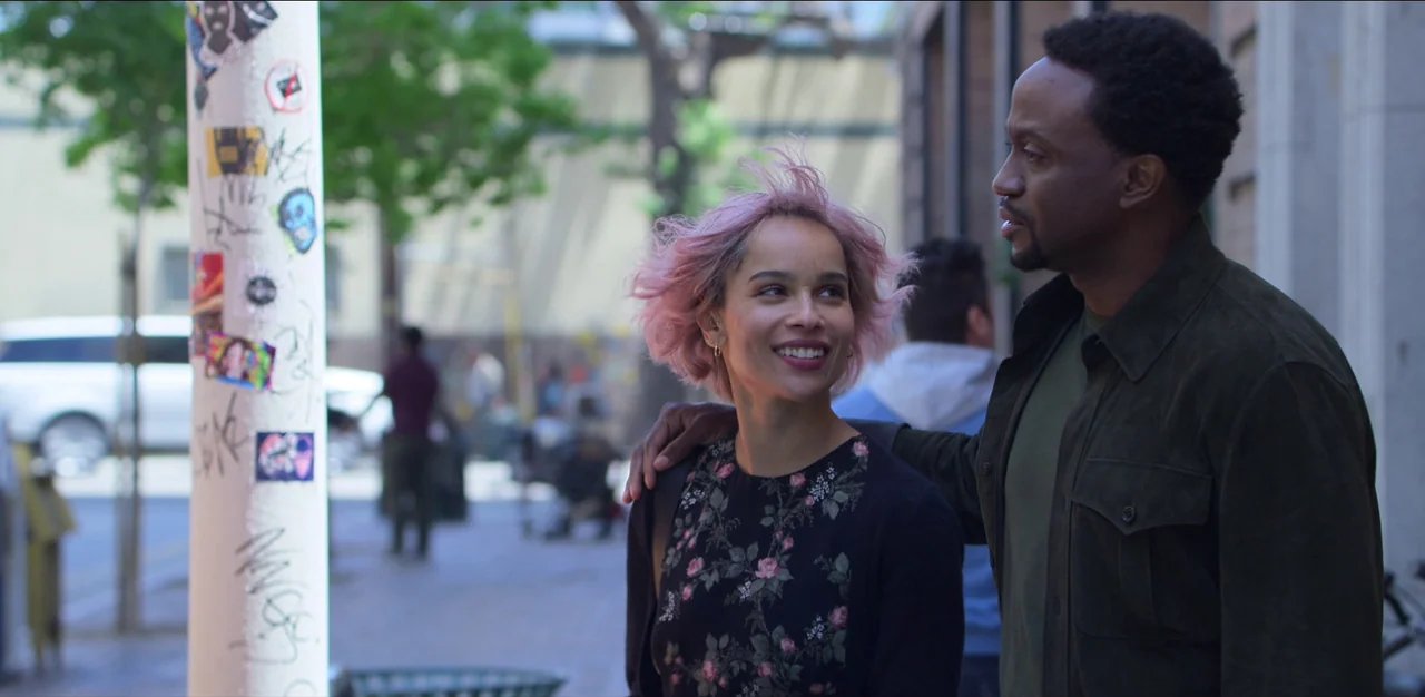 Kimi (2022) movie still: medium — An urban sidewalk features a white post plastered with various colorful stickers and graffiti. A man and woman…