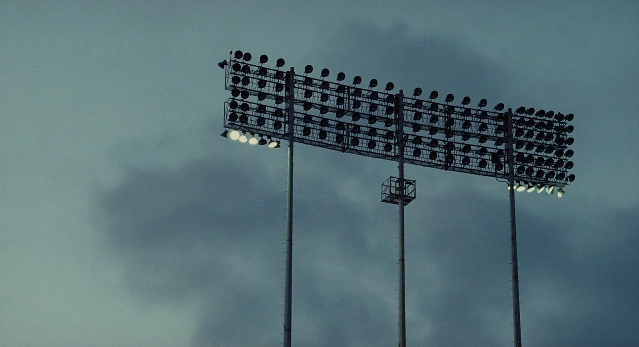 Moneyball (2011) movie still: establishing — A tall metal structure supports rows of stadium floodlights against a cloudy evening sky. The metal…