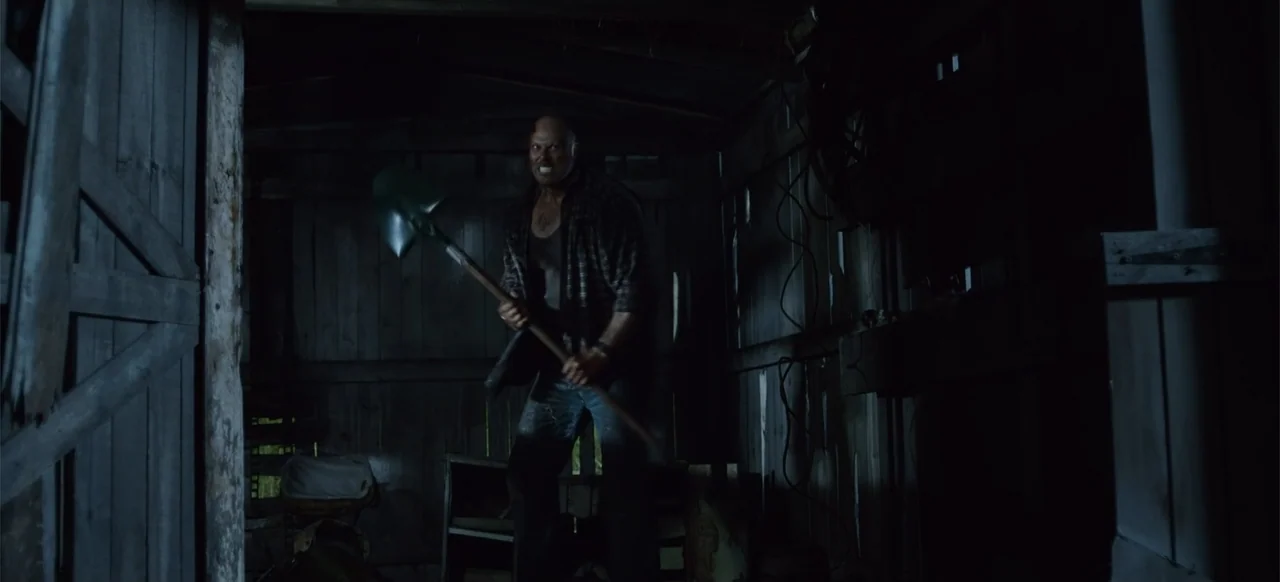 Boar (2018) movie still: medium — A dark wooden interior features rough timber walls and clutter in the low light. A man standing in the doorway…