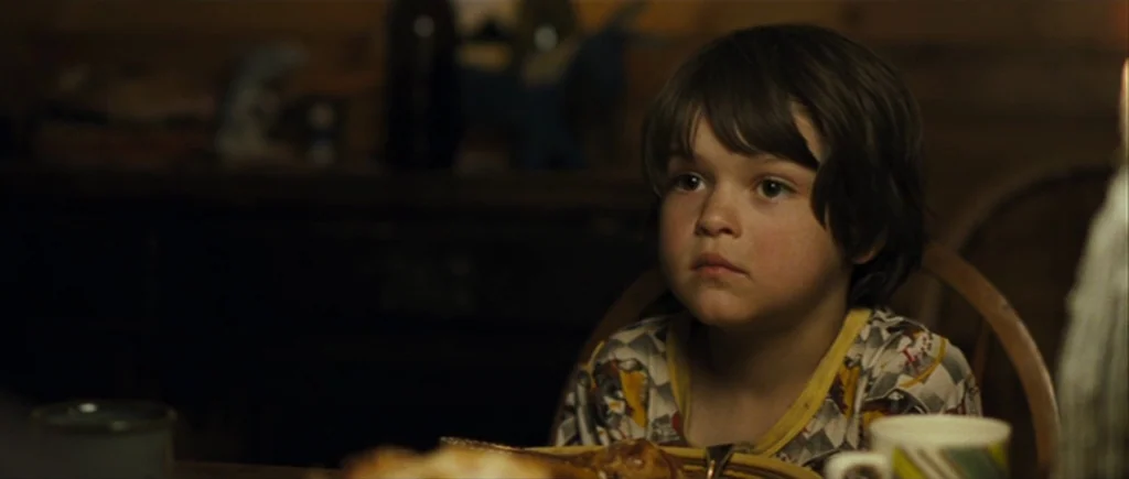 The Hunter (2011) movie still: closeup — A dimly lit room features a wooden table and a child sitting in a chair. The boy looks steadily ahead while…