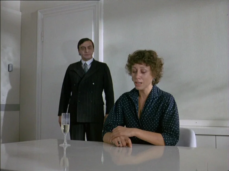 Satan’s Brew (1976) movie still: medium — A spartan room features white walls and a reflective white tabletop. A woman sits while a man stands…