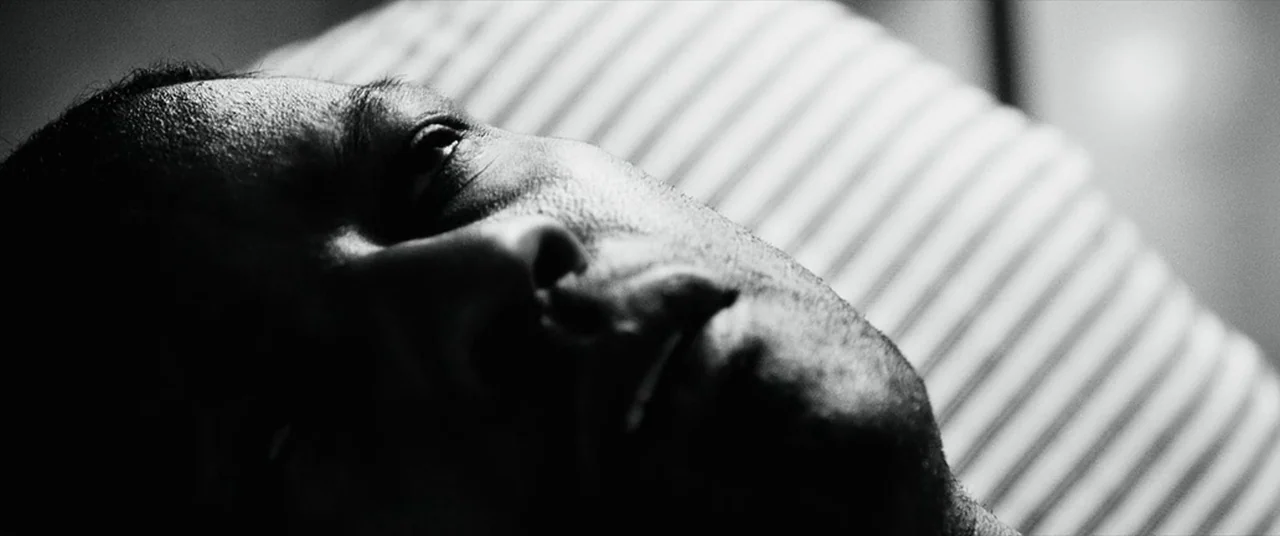Man on Fire (2004) movie still: closeup — A man lies on a striped fabric surface under low-key light. His face is angled toward the top of the frame…