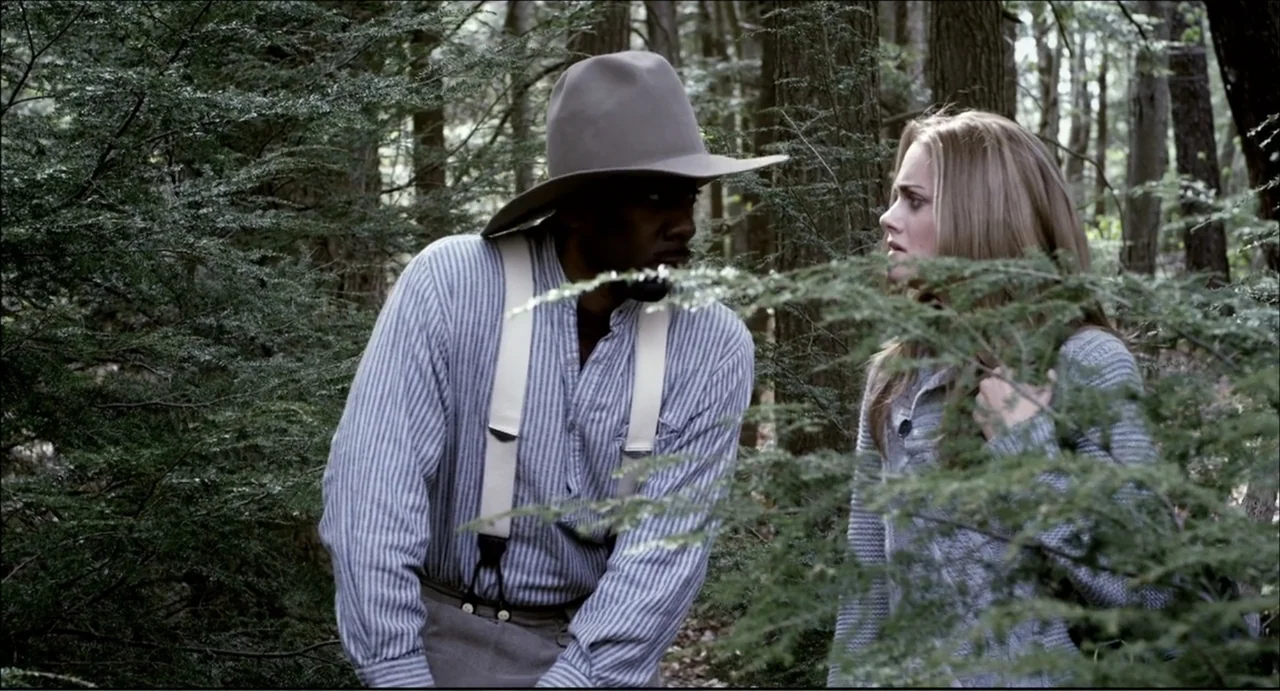 The Secret Village (2013) movie still: medium — A dense patch of pine trees forms a barrier around two individuals standing together. A man in a…