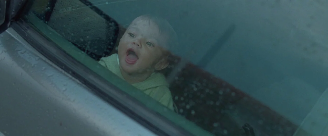 The End We Start From (2023) movie still: closeup — A toddler is viewed through the wet, rain-streaked glass of a car window. The child looks…