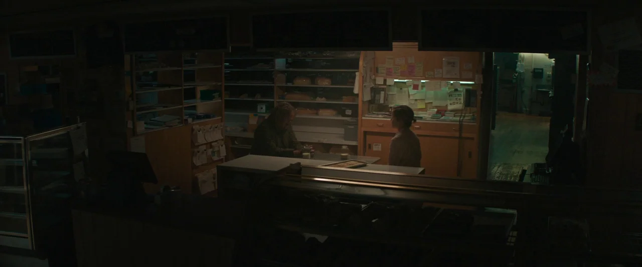 Pig (2021) movie still: wide — The interior of a dim bakery at night features dark wooden shelves and counters. Two people sit across from each…