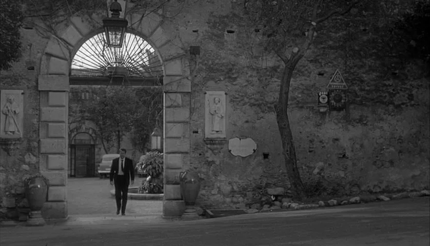 L’Avventura (1960) movie still: wide — A stone courtyard is enclosed by weathered walls featuring arched entryways and carved reliefs. A man in a…