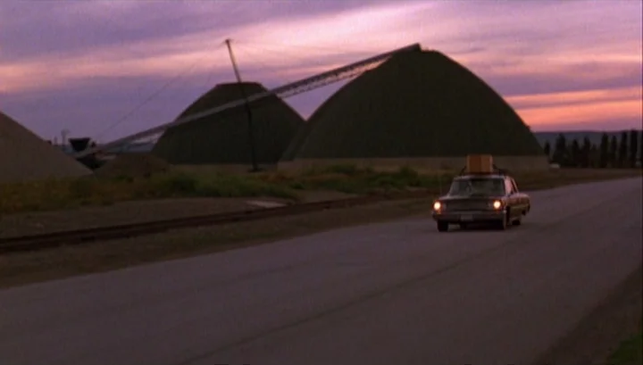 Highway 61 (1991) movie still: wide — A rural highway stretches past large conical industrial storage mounds under a sunset sky. A vintage car with…