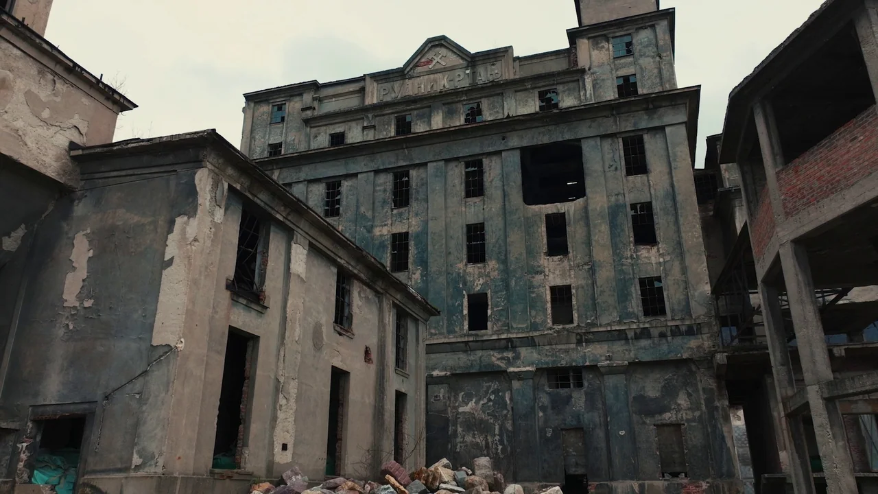 Helll Hole (2024) movie still: establishing — A multi-story abandoned industrial building with weathered concrete walls and numerous shattered…