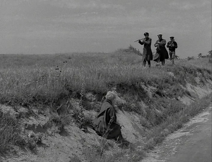 La Strada (1954) movie still: wide — An open rural landscape features a steep dirt embankment covered in tall grasses. A lone person sits on the…