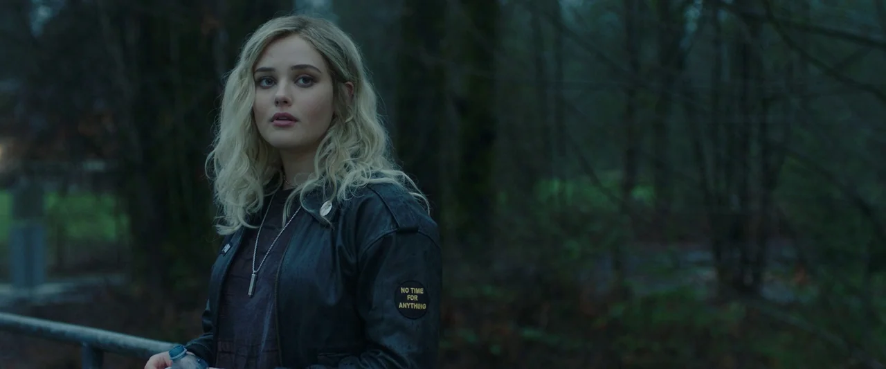 Spontaneous (2020) movie still: medium — A woman stands in a dense, overcast woodland setting with bare trees and greenery. The subject looks…