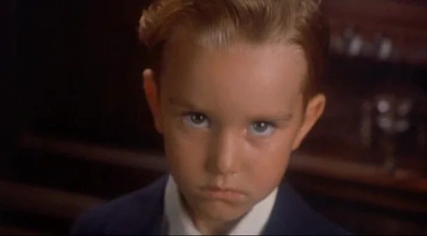 Boxing Helena (1993) movie still: closeup — A dimly lit interior space features dark, indistinct surfaces behind a young boy. The boy stares…