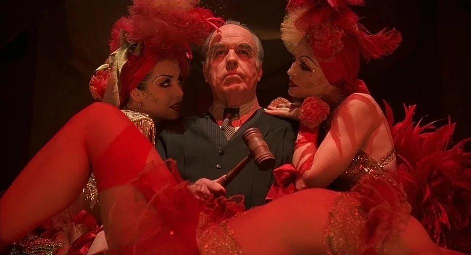 Chicago (2002) movie still: medium — A dark cabaret setting features an elderly man in a suit surrounded by showgirls. The man grips a wooden gavel…
