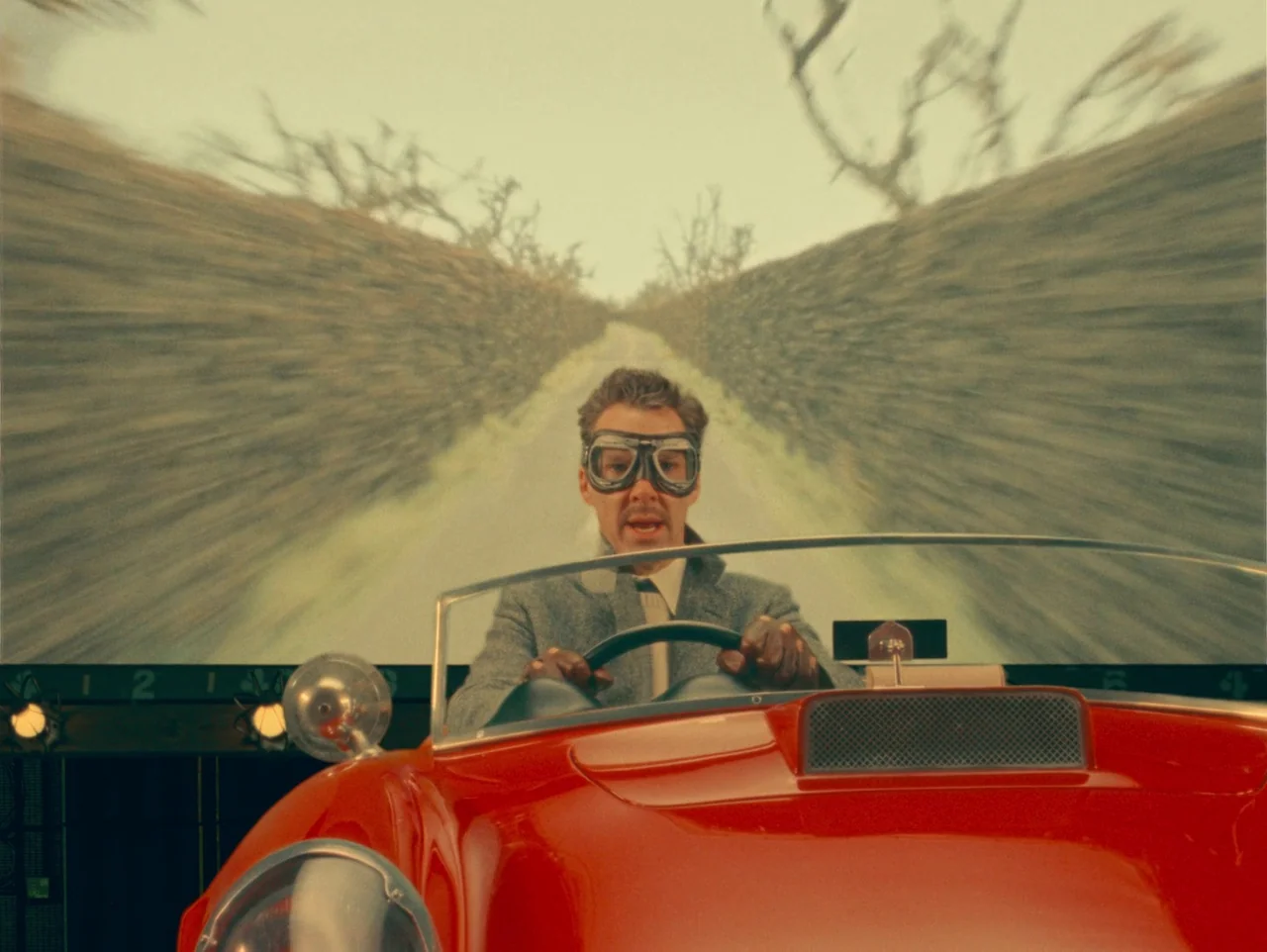 The Wonderful Story of Henry Sugar (2023) movie still: medium — A man sits in a bright red vintage convertible on a studio set. A rear projection…