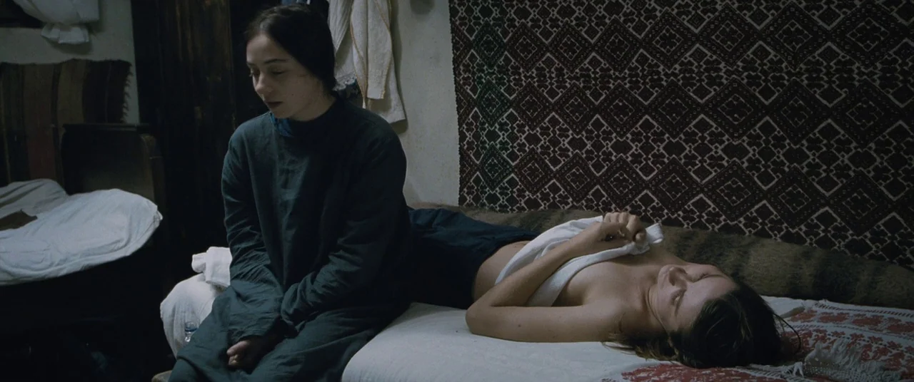 Beyond the Hills (2012) movie still: medium — A dimly lit room features a patterned textile hung on the wall. A woman sits on the edge of the bed as…