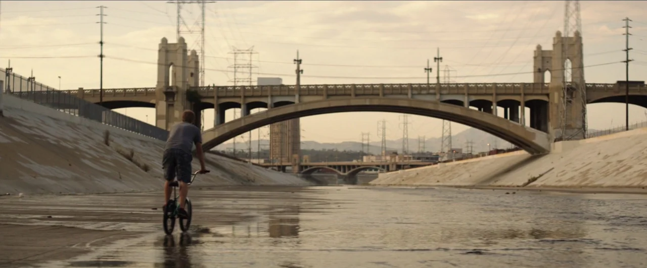 Dean (2016) movie still: wide — A wide concrete channel features high, sloped walls and overhead power lines stretching across. A lone teenager…