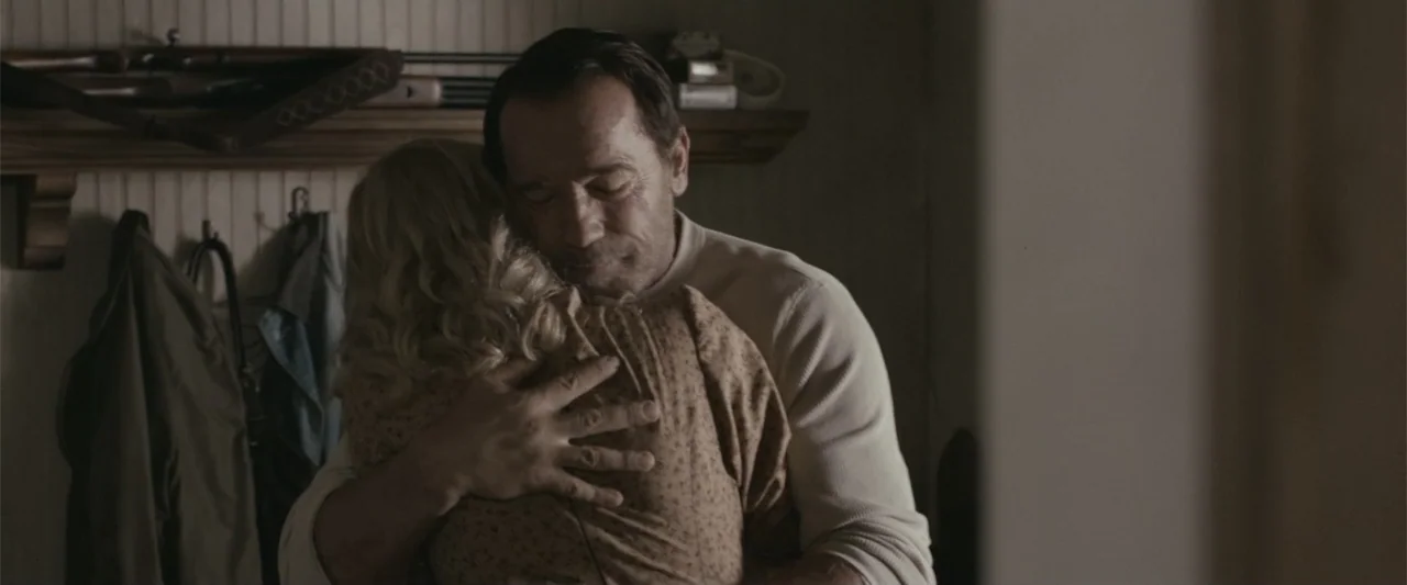 Maggie (2015) movie still: medium — A rural interior room with beadboard walls and a wooden shelf holding a shotgun. An adult man hugs a blonde…