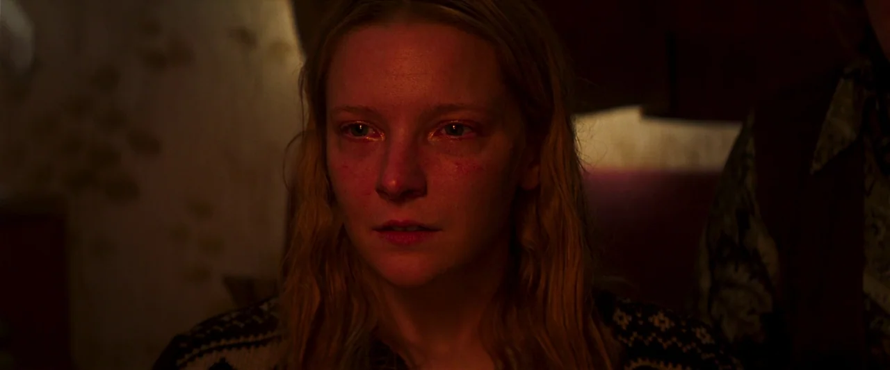 Starve Acre (2023) movie still: closeup — A dimly lit indoor space with low ambient light level. A young woman with blonde hair stares directly…