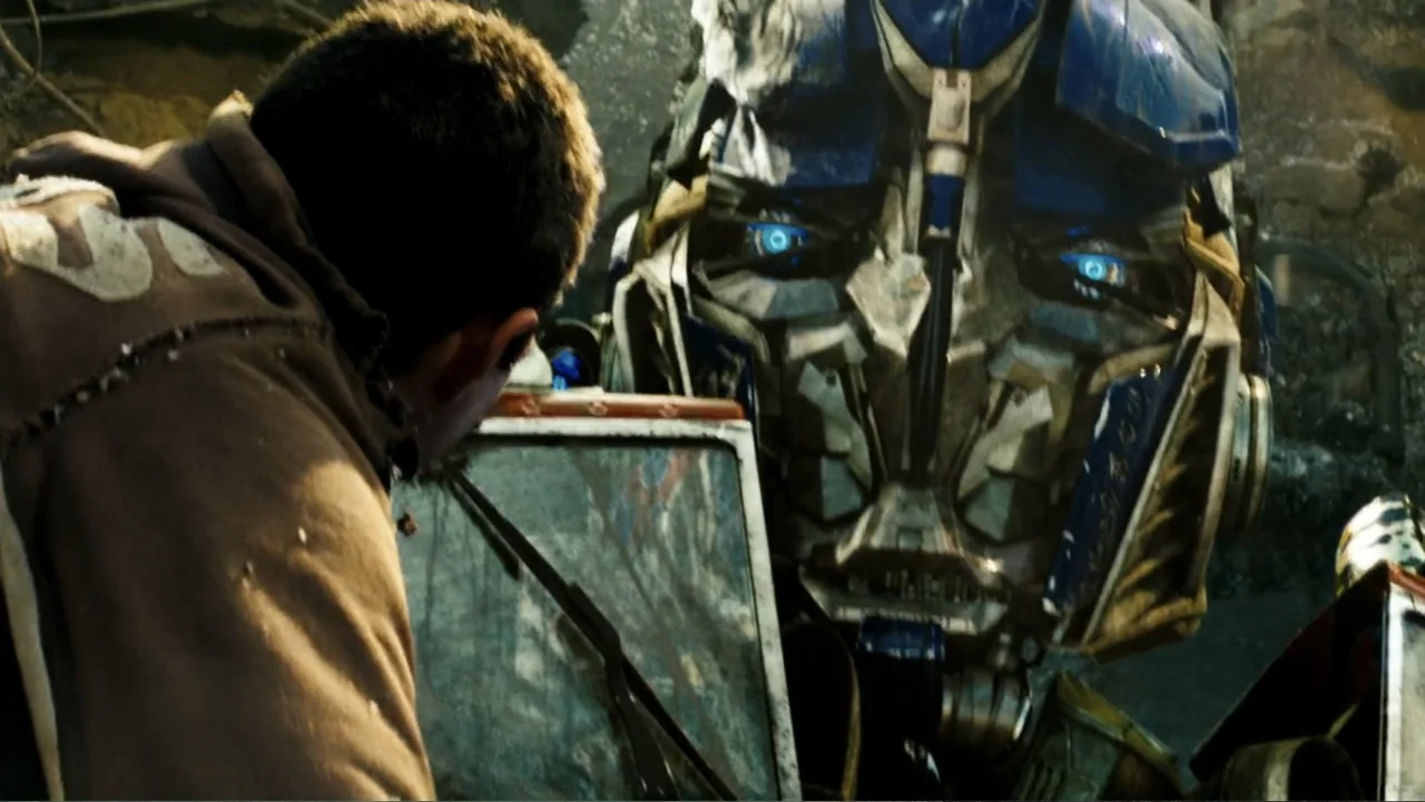 Transformers (2007) movie still: medium — A debris-covered outdoor area with broken metallic structures and rubble in the background. A young man in…