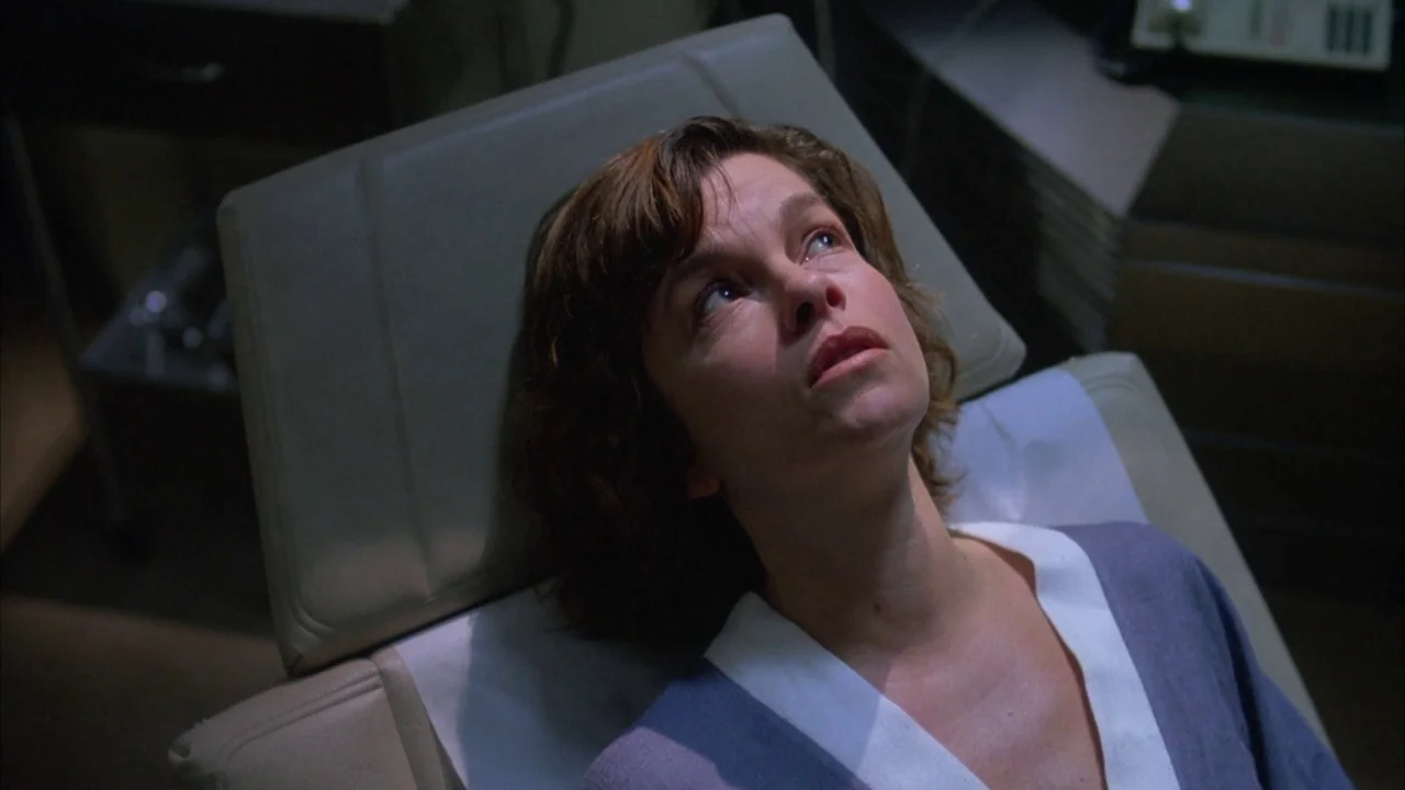 Dead Ringers (1988) movie still: closeup — A dimly lit medical examination room contains a reclining chair and basic furniture. A woman in a…