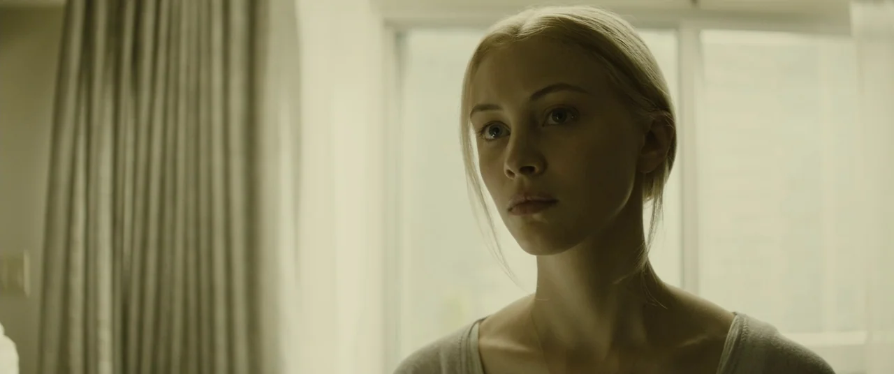 Enemy (2013) movie still: closeup — A room features a large window covered by light curtains. A young woman looks toward the side while standing in…