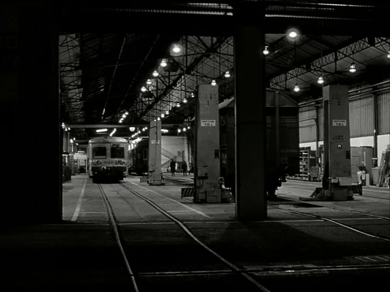 Eloge de L’amour (2001) movie still: establishing — A vast, cavernous railway maintenance hangar features concrete pillars and multiple sets of…