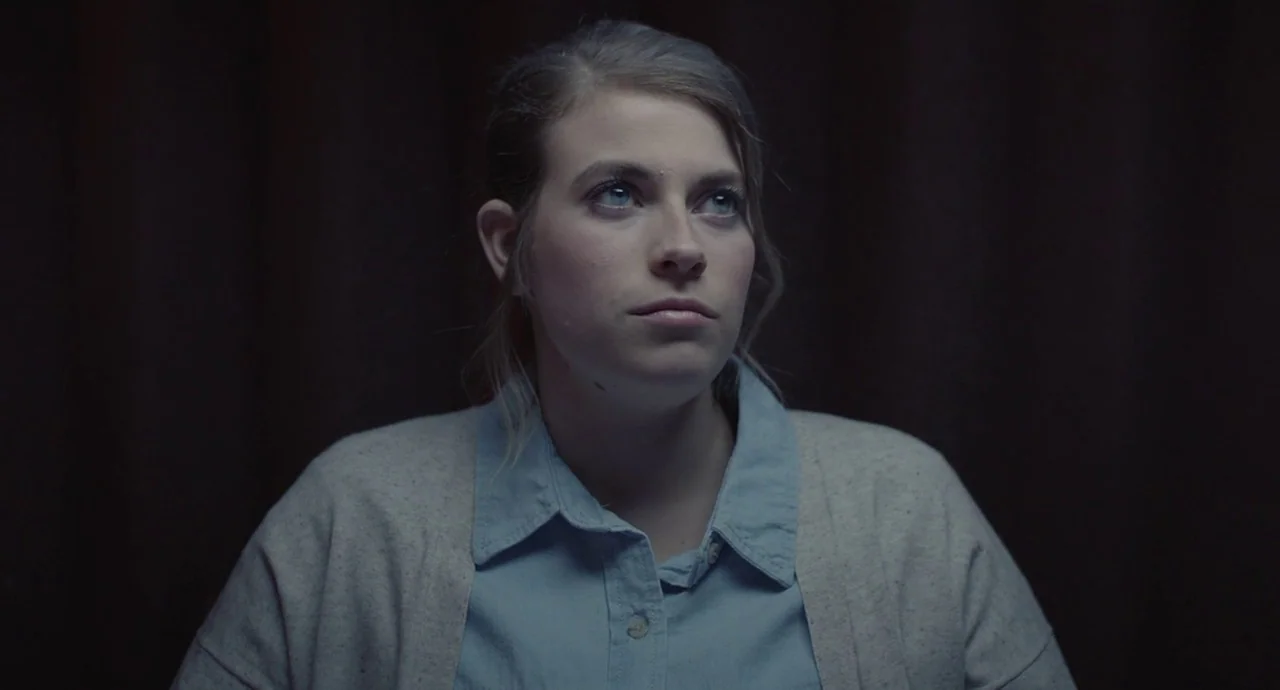 The Wolf of Snow Hollow (2020) movie still: medium — A woman stands against a dark, featureless backdrop in an interior setting. She gazes upward…