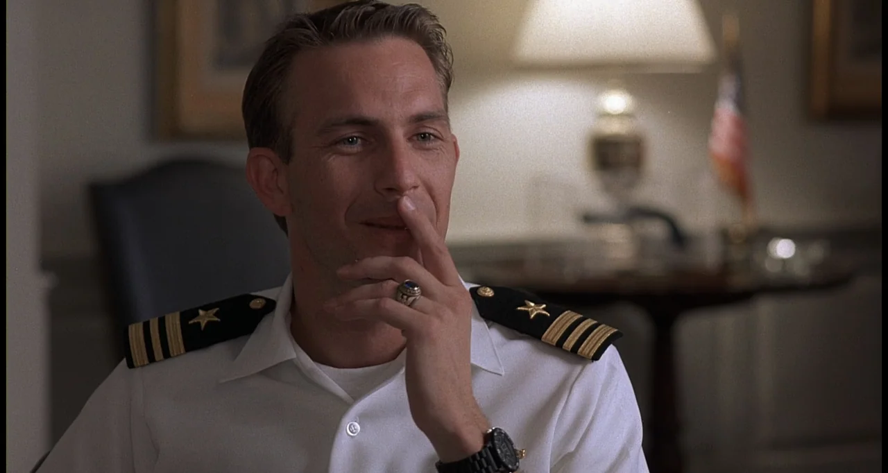 No Way Out (1987) movie still: medium — A well-lit office interior features a wooden table and a blurred American flag background. A man in a naval…