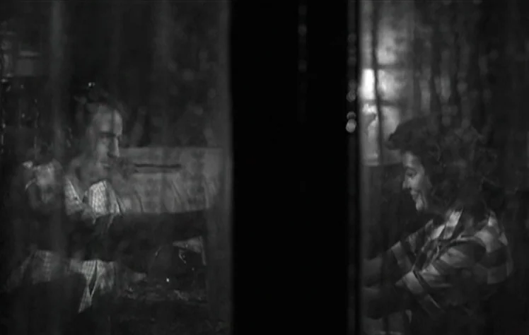 On The Beach (1959) movie still: medium — Two individuals stand facing each other separated by a dark glass vertical partition. The subjects appear…