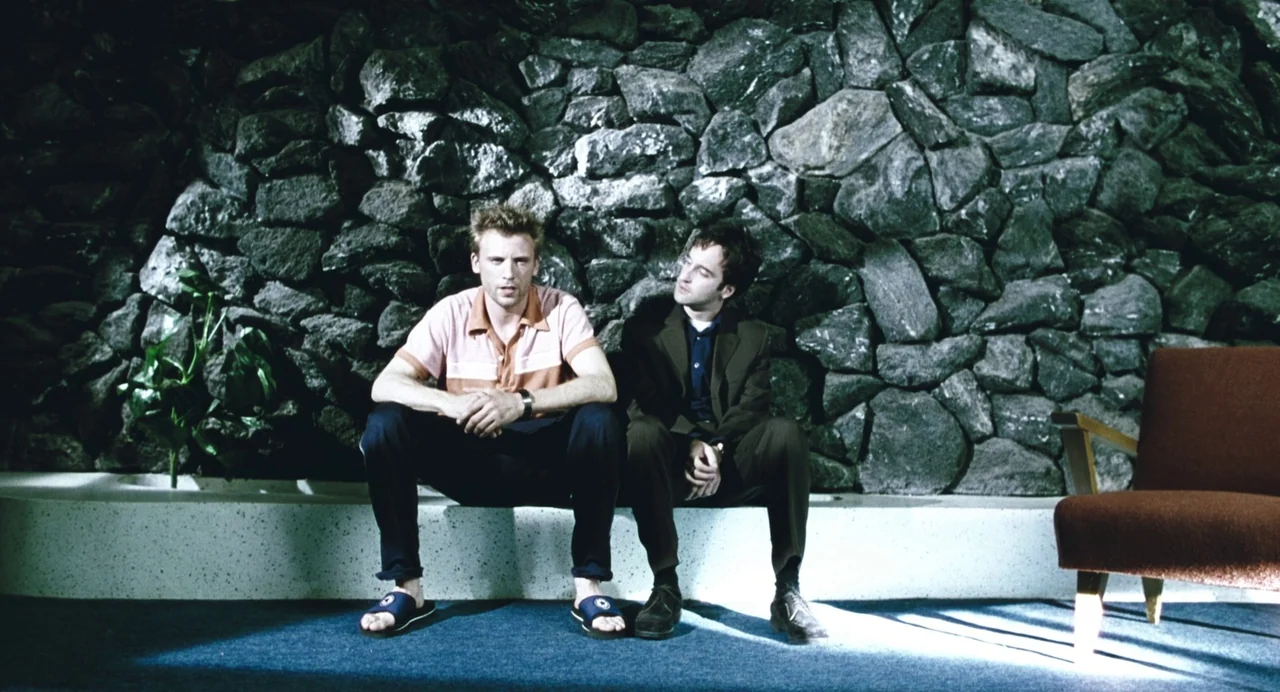 Last Night (1998) movie still: fullbody — A room with a rough stone wall and a raised white concrete bench. Two men sit on the bench while one looks…