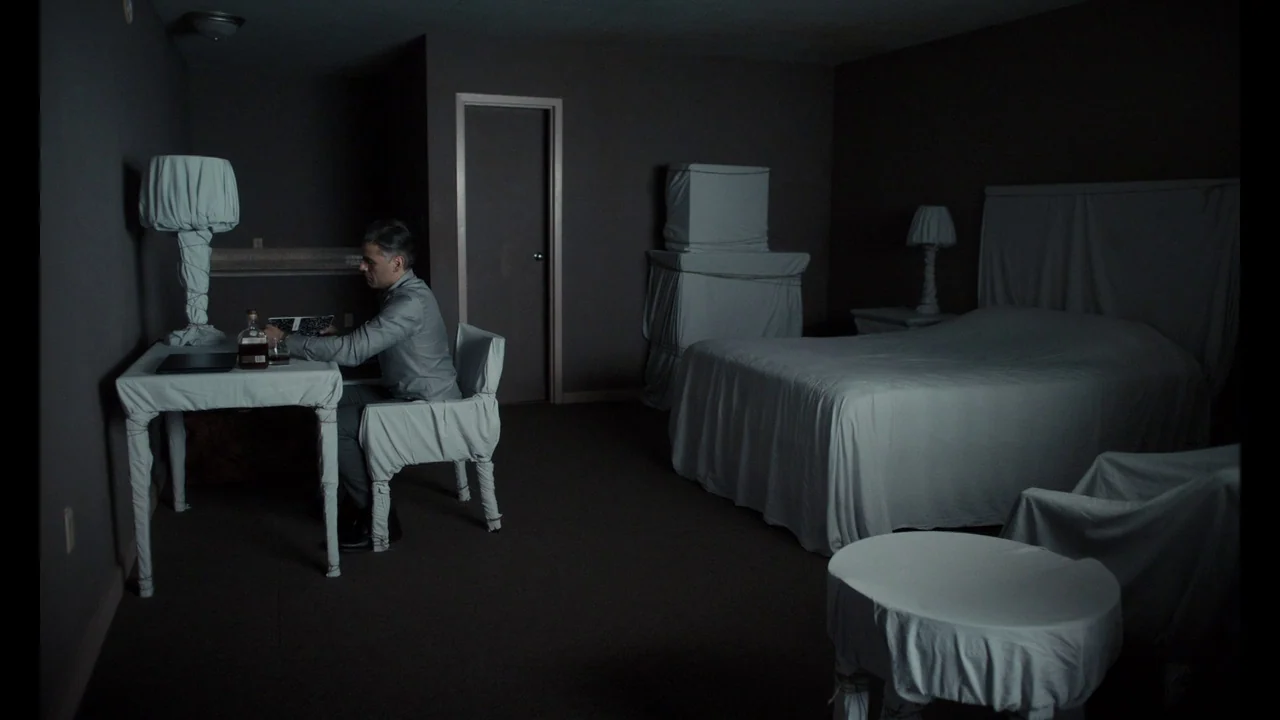 The Card Counter (2021) movie still: wide — A dark hotel room features furniture draped entirely in white fabric sheets. A man sits at a desk in the…