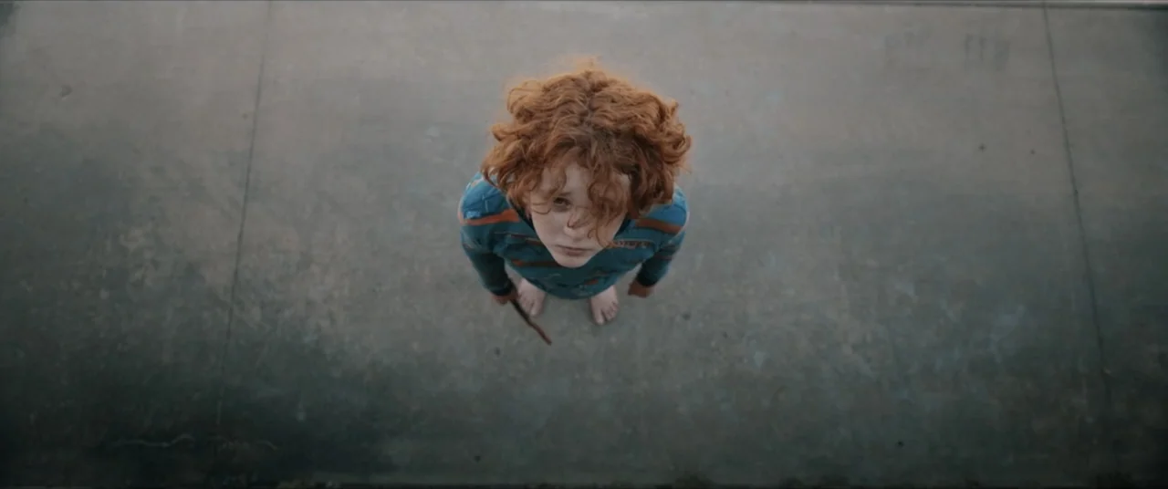 Possessions (2024) movie still: fullbody — A child stands alone on a grey concrete surface viewed from directly above. The boy with curly ginger…