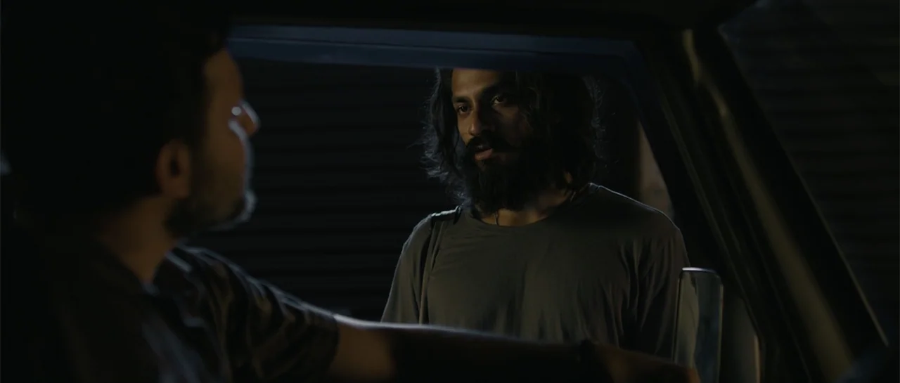 Nishaanchi 2 (2025) movie still: medium — A man with long hair and a beard stands outside a parked vehicle at night. He is talking to another man…