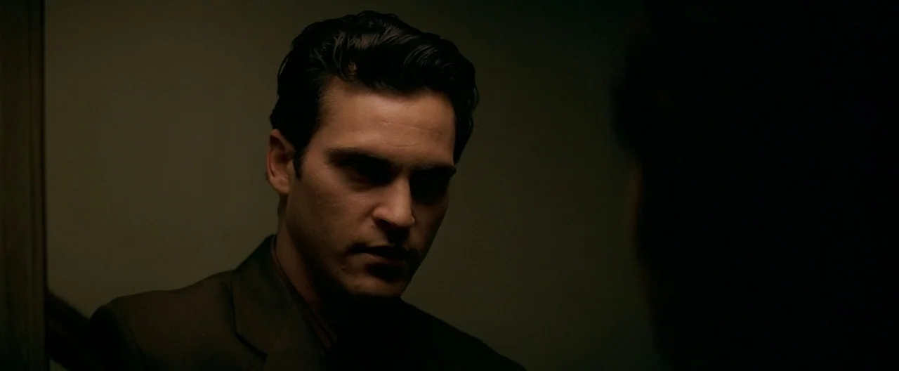 The Yards (2000) movie still: closeup — A dark, dimly lit indoor setting with shadowy walls and minimal visible detail. A man in a dark suit gazes…