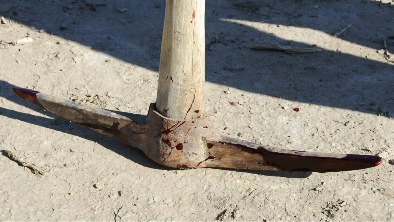 Savageland (2015) movie still: closeup — A rusted metal pickaxe rests horizontally on cracked, sun-baked dry dirt. Bloodstains smear the head of the…