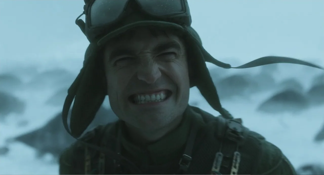 Mickey 17 (2025) movie still: closeup — A desolate, snowy mountain peak under overcast skies. A man wearing military gear and goggles braces against…