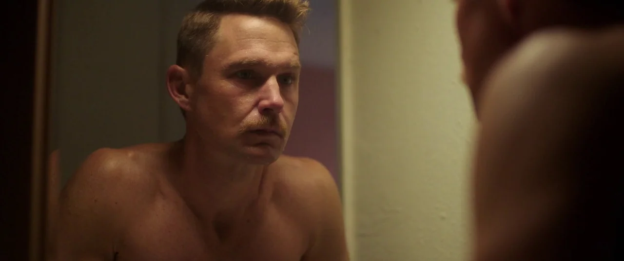 Blindfire (2020) movie still: closeup — A man stands in front of a mirror in a dimly lit room. He looks directly at his own reflection with a steady…
